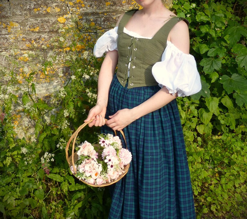 May include: A woman wearing a green and white dress with a basket of pink and white flowers. The dress has a green bodice with a lace-up front and a plaid skirt.