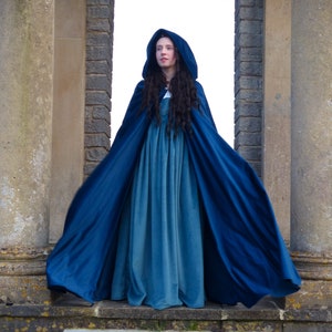 May include: A woman wearing a long, flowing blue hooded cloak. The cloak is made of a soft, velvety fabric and has a deep blue color. The woman is standing in front of a stone archway.