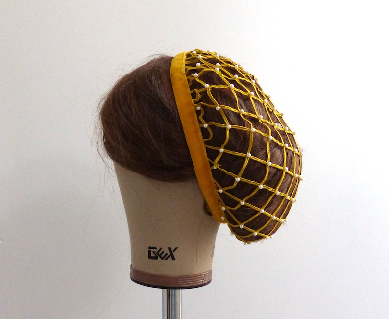 以下が含まれることがあります： 白いビーズが付いた黄色のヘアネット。ヘアネットは細かいメッシュ素材でできており、ベルベットのバンドが付いています。