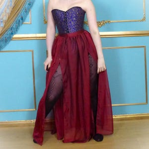 May include: A long, burgundy skirt with two slits at the front. The skirt is made of a sheer fabric and is worn over a purple and gold brocade corset.