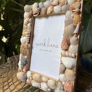 May include: A rectangular photo frame decorated with various seashells. The frame is made of light-colored wood and features the text "park lane PHOTO FRAME | 4X6". The seashells are in shades of white, tan, and brown, with small pearl accents.