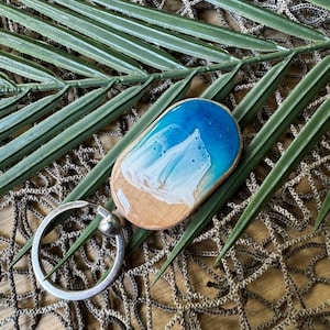 May include: Wooden keychain with a beach scene design. The keychain features a silver metal ring and a wooden oval pendant with a blue and white ocean wave design over a tan beach. The keychain is displayed on a background of green leaves and a woven net.