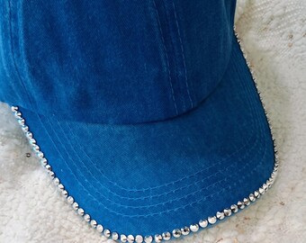 May include: A blue baseball cap with a curved peak. The peak is edged with small, shiny, silver-coloured rhinestones. The cap is made of a soft, textured fabric.