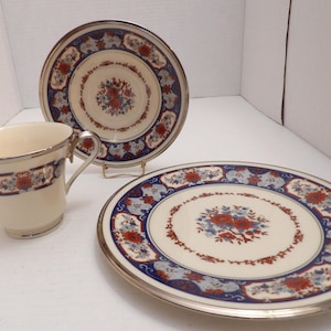 May include: A cream-colored teacup, saucer, and plate set. The set features a floral design with red and blue accents on a white background, and a silver trim. The plate is approximately 10 inches in diameter.