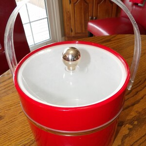 May include: A vintage red ice bucket with a clear plastic handle. The bucket has a white interior and a gold-colored knob on the lid. The handle arches over the top, and the bucket sits on a wooden surface.