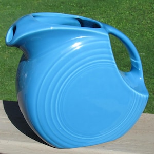 May include: A turquoise-blue ceramic pitcher with a large handle and a spout. The pitcher has a rounded body with concentric, ridged detailing. The handle is a curved, open design. The pitcher is likely vintage and suitable for serving beverages.