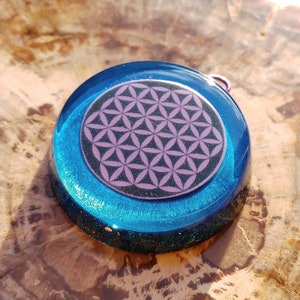May include: A round, blue resin pendant with a purple flower of life design. The pendant is on a wooden surface.