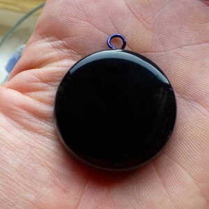 Puede incluir: Un colgante redondo negro brillante con un pequeño lazo azul. El colgante se muestra en una mano abierta, resaltando su superficie lisa y reflectante. La forma circular del colgante es prominente.