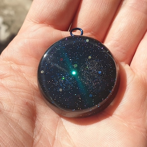 May include: A round, black resin pendant with a galaxy design. The pendant features iridescent glitter and a green nebula in the center.