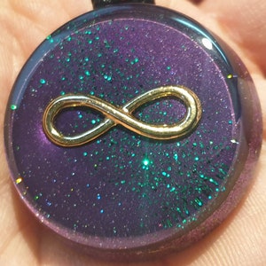 May include: A round pendant with a gold infinity symbol on a purple background with green glitter.