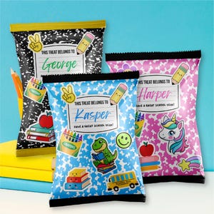 Puede incluir: Tres bolsas de snacks rectangulares con diseños escolares. La bolsa negra dice George, la azul Kasper y la rosa Harper. Cada bolsa presenta ilustraciones de crayones, lápices, libros y un unicornio o un dinosaurio.