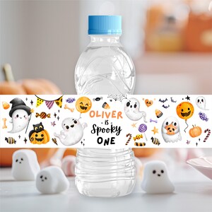 May include: A clear plastic water bottle with a light blue cap, featuring a white label decorated with watercolor illustrations of ghosts, pumpkins, candy, and bats. The text on the label reads, "OLIVER is SPOOKY ONE" in orange and black lettering.
