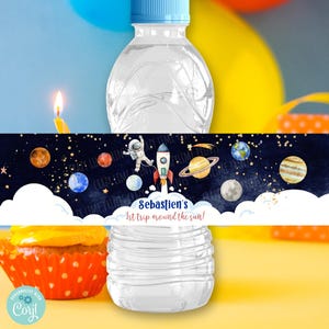 Puede incluir: Etiqueta de botella de agua con temática espacial. La etiqueta presenta un fondo azul con estrellas, planetas y un cohete. El texto en la etiqueta dice "Sebastien's 1st trip around the sun!"