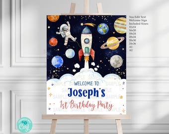 May include: A welcome sign for a 1st birthday party with a space theme. The sign features a rocket, an astronaut, planets, and stars on a dark blue background. The text reads "Welcome to Joseph's 1st Birthday Party".