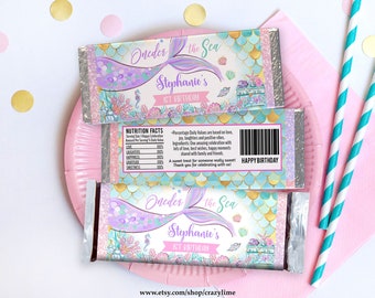 May include: Chocolate bar wrappers with a mermaid theme, featuring a purple and teal color scheme. The text reads "Oneder the Sea" and "Stephanie's 1st Birthday." The wrappers are decorated with mermaid tails, seashells, and a cake.