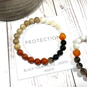 May include: Two beaded bracelets with a variety of stones, including brown, white, black, and orange. The bracelets are on a white wooden surface. The text on the card reads "Crystal Jewellery Protection Black Tourmaline - Jasper - Agate".