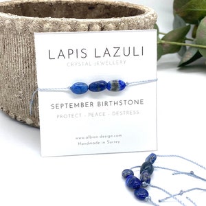 May include: A blue lapis lazuli gemstone bracelet with a light blue cord. The card reads "Lapis Lazuli Crystal Jewellery September Birthstone Protect Peace Destress www.albion-design.com Handmade in Surrey".