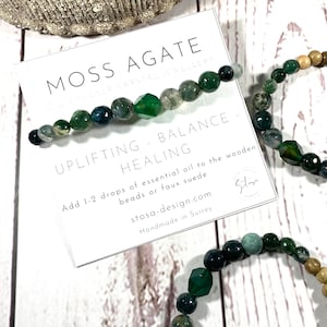 Op de afbeelding: Moosagaten armband met groene en bruine kralen. De armband ligt op een wit houten oppervlak met een kaart waarop staat: "Moss Agate - Uplifting - Balance - Healing. Add 1-2 drops of essential oil to the wooden beads or faux suede. stosa-design.com Handmade in Surrey."