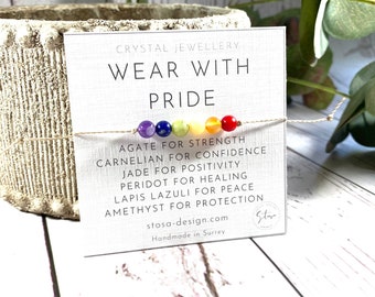 May include: A rainbow colored bracelet made with agate, carnelian, jade, peridot, lapis lazuli, and amethyst stones. The bracelet is on a white card with the text "CRYSTAL JEWELLERY WEAR WITH PRIDE AGATE FOR STRENGTH CARNELIAN FOR CONFIDENCE JADE FOR POSITIVITY PERIDOT FOR HEALING LAPIS LAZULI FOR PEACE AMETHYST FOR PROTECTION stosa-design.com Handmade in Surrey Star".
