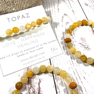 Pu&ograve; includere: Due braccialetti realizzati con perline di topazio. I braccialetti sono gialli e bianchi e hanno un accento di perline di legno. I braccialetti sono su una superficie di legno bianco con un biglietto che dice "Topazio Amore - Felicit&agrave; - Guarigione Aggiungi 1-2 gocce di olio essenziale alle perline di legno o al camoscio sintetico stosa-design.com Fatto a mano a Surrey"