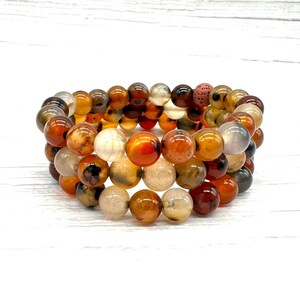 May include: Three bracelets composed of polished, round beads in shades of orange, brown, cream, and grey. The bracelets are stacked and presented on a white surface.