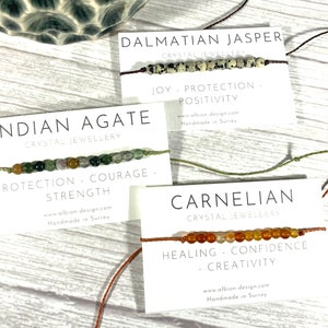 Op de afbeelding: Drie armbanden gemaakt met verschillende soorten edelstenen. De armbanden liggen op witte kaartjes met de namen van de stenen, "Dalmatische Jaspis", "Indiase Agate" en "Carneol". Elk kaartje vermeldt ook de eigenschappen van de steen, zoals "Vreugde - Bescherming - Positiviteit", "Bescherming - Moed - Kracht" en "Genezing - Zelfvertrouwen - Creativiteit".