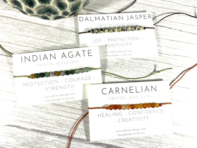Op de afbeelding: Drie armbanden gemaakt met verschillende soorten edelstenen. De armbanden liggen op witte kaartjes met de namen van de stenen, "Dalmatische Jaspis", "Indiase Agate" en "Carneol". Elk kaartje vermeldt ook de eigenschappen van de steen, zoals "Vreugde - Bescherming - Positiviteit", "Bescherming - Moed - Kracht" en "Genezing - Zelfvertrouwen - Creativiteit".