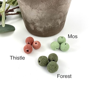 May include: Four red lava rock beads labeled "Thistle" and four green lava rock beads labeled "Mos" and "Forest" on a white surface.