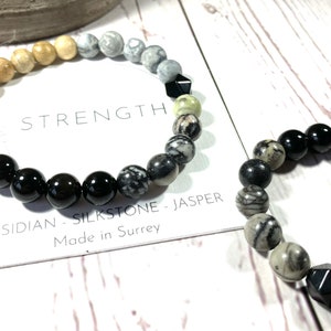 May include: Two beaded bracelets, one with brown, grey, and black beads, and the other with black and grey beads. The bracelet with brown, grey, and black beads has the text "STRENGTH OBSIDIAN - SILKSTONE - JASPER Made in Surrey" on a white card.