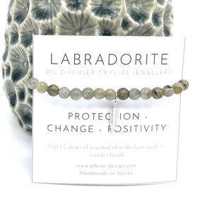 May include: A Labradorite oil diffuser crystal jewellery bracelet. The bracelet has small, round, grey-green beads and a silver feather charm. The card behind the bracelet reads "Protection - Change - Positivity."