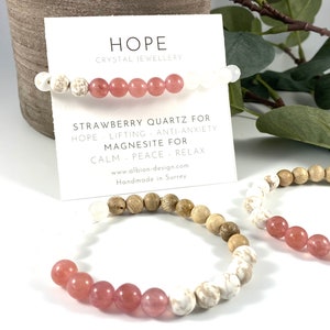 May include: Three bracelets made with natural stones. The first bracelet is made with white and pink beads. The second bracelet is made with brown wood beads. The third bracelet is made with white and pink beads. The card reads "HOPE Crystal Jewellery Strawberry Quartz for Hope Lifting Anti-Anxiety Magnesite for Calm Peace Relax www.albion-design.com Handmade in Surrey"