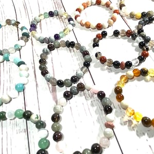 May include: A collection of colourful beaded bracelets made with various stones and crystals. The bracelets are arranged on a white wooden surface.
