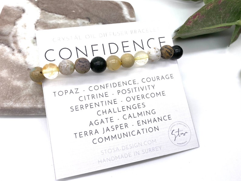 May include: A bracelet made with various semi-precious stones, including topaz, citrine, serpentine, agate, and terra jasper. The bracelet is on a white card with the word "CONFIDENCE" in black lettering and a description of the stones and their meanings.