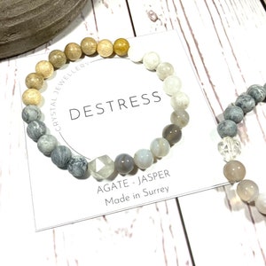 May include: A grey, white and brown beaded bracelet with a clear crystal bead. The bracelet is on a white card with the text "DESTRESS" and "AGATE - JASPER Made in Surrey".