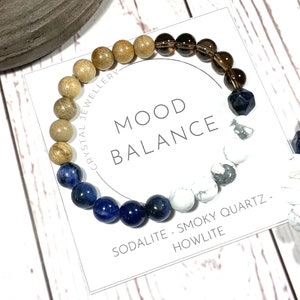 Peut inclure: Bracelet "Équilibre de l'humeur" avec des perles en bois brun, sodalite bleue, howlite blanche et quartz fumé. Le bracelet est présenté sur une carte blanche avec le texte "Équilibre de l'humeur" et "Sodalite - Quartz fumé - Howlite".