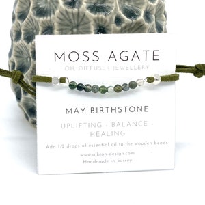 May include: Green moss agate gemstone bracelet with silver beads and green suede cord. The bracelet is labelled "Moss Agate Oil Diffuser Jewellery" and "May Birthstone". The card also says "Uplifting - Balance - Healing" and "Add 1-2 drops of essential oil to the wooden beads".