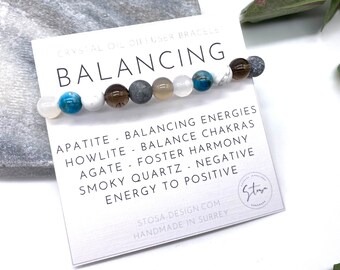 May include: A white card with the text "CRYSTAL OIL DIFFUSER BRACELET BALANCING" and a list of stones and their properties. The card is next to a bracelet made of various stones, including white, gray, brown, and blue.