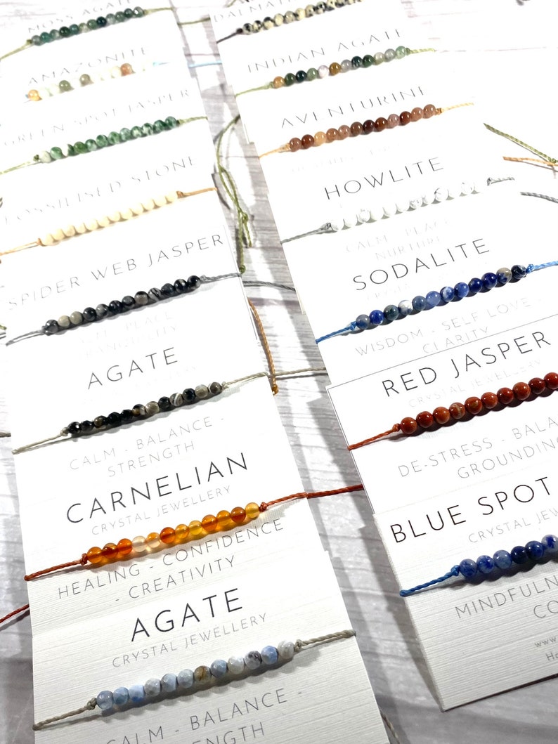 Op de afbeelding: Een verzameling kleurrijke armbanden gemaakt van verschillende soorten edelstenen. Elke armband heeft een kaart met de naam van de steen en de bijbehorende helende eigenschappen. De stenen omvatten agaat, aventurijn, howliet, sodaliet, rode jaspis, carneol en blauwe vlek.