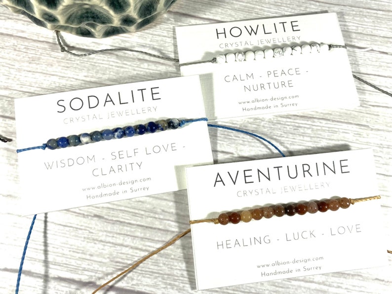 Op de afbeelding: Drie armbanden gemaakt met verschillende soorten kristallen. De armbanden liggen op witte kaartjes met de naam van het kristal, de eigenschappen van het kristal en de website en locatie van de maker. De kristallen zijn howliet, sodaliet en aventurijn.