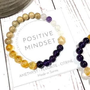 May include: A bracelet made with amethyst, fluorite, citrine, and wood beads. The bracelet is on a white card with the text "POSITIVE MINDSET" and "AMETHYST - FLUORITE - CITRINE" and "Made in Surrey".
