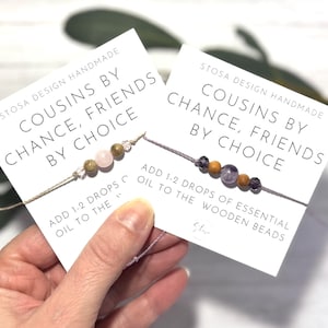 Pode incluir: Dois cartões brancos com o texto "Cousins by Chance, Friends by Choice" e instruções para adicionar 1-2 gotas de óleo essencial às contas de madeira. Cada cartão tem um bracelete de corda com contas de madeira e pedras preciosas.