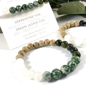 Puede incluir: Dos pulseras hechas con cuentas de serpentina verde y jaspe verde, con cuentas de madera y cuentas blancas. Las pulseras están sobre una superficie blanca. El texto en la tarjeta dice: "Serpentina para la positividad - Limpiar + Curar Jaspe verde para la alegría - Positividad + Calma www.albion-design.com Hecho a mano en Surrey"