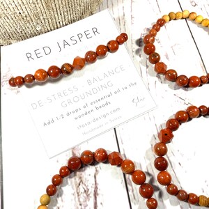 以下が含まれることがあります： 赤いジャスパーと木製のビーズのブレスレット。カードには「Red Jasper Grounding De-Stress. Balance. Add 1-2 drops of essential oil to the wooden beads stosa-design.com Handmade in Surrey」と書かれています。