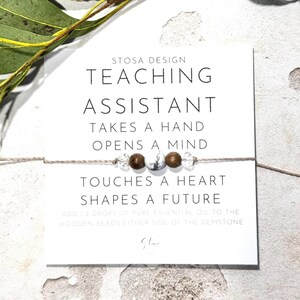 May include: A white card with the text "STOSA DESIGN TEACHING ASSISTANT TAKES A HAND OPENS A MIND TOUCHES A HEART SHAPES A FUTURE" and a small string bracelet with brown and white beads.