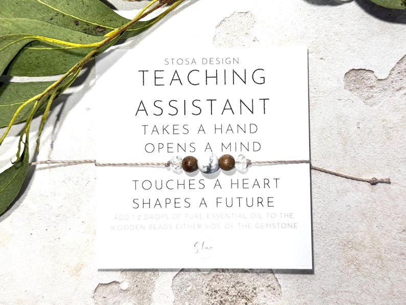 May include: A white card with the text "STOSA DESIGN TEACHING ASSISTANT TAKES A HAND OPENS A MIND TOUCHES A HEART SHAPES A FUTURE" and a small string bracelet with brown and white beads.
