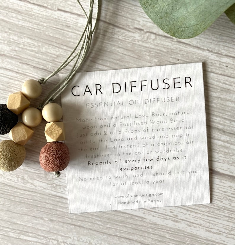 May include: A car diffuser made from natural lava rock, wood, and a fossilized wood bead. The diffuser is a necklace with beads in shades of brown, beige, and black. The card reads "CAR DIFFUSER ESSENTIAL OIL DIFFUSER Made from natural Lava Rock, natural wood and a Fossilised Wood Bead Just add 2 or 3 drops of pure essential oil to the Lova and wood and pop in the car. Use instead of a chemical air freshener in the car or wardrobe. Reapply oil every few days as it evaporates. No need to wash, and it should last you for at least a year. www.albion-design.com Handmade in Surrey"