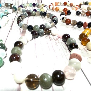 May include: A collection of colorful beaded bracelets made with various stones, including white, green, brown, black, and orange. The bracelets are arranged in a circle on a white wooden surface.