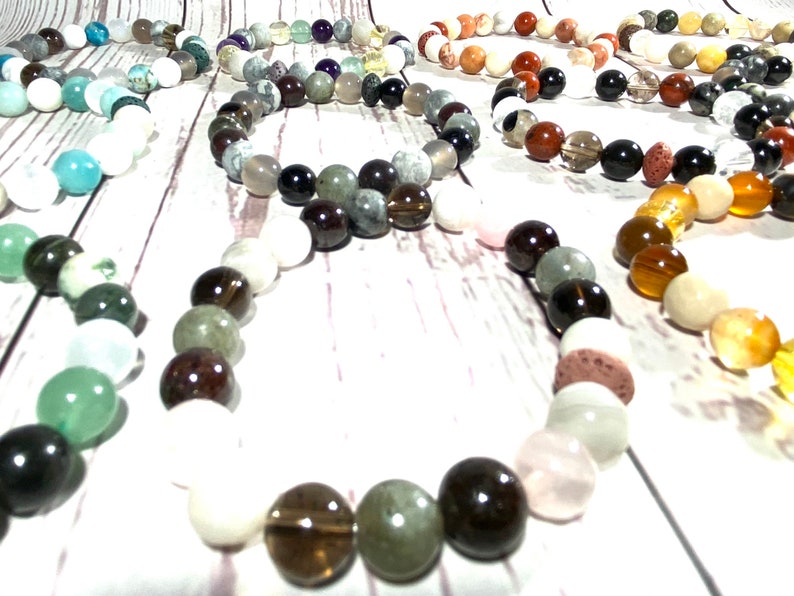 May include: A collection of colorful beaded bracelets made with various stones, including white, green, brown, black, and orange. The bracelets are arranged in a circle on a white wooden surface.