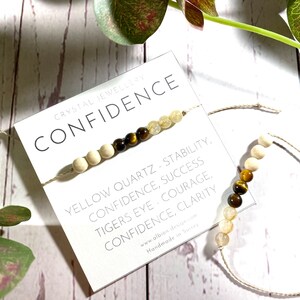 May include: Two bracelets made with natural stones and string. The bracelets are on a white wooden surface. The top bracelet has yellow quartz, tiger's eye, and wood beads. The bottom bracelet has tiger's eye and yellow quartz beads. A card with the text "CRYSTAL JEWELLERY CONFIDENCE YELLOW QUARTZ - STABILITY, CONFIDENCE, SUCCESS TIGERS EYE - COURAGE, CONFIDENCE, CLARITY www.albion design.com Handmade in Sussex" is visible in the image.