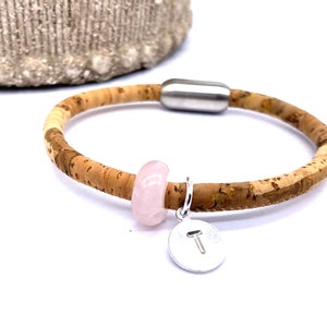 May include: A cork bracelet with a pink stone charm and a silver charm with the letter "T" on it.